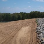 A sloped area with freshly leveled brown soil from recent excavating shows tire tracks on the left, and a line of large gray rocks on the right, bordered by green trees under a blue sky. Kentucky Builders And Excavating
