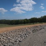 A dirt road curves through a grassy field bordered by a rocky embankment, where excavating work hints at building a new pole barn amid green trees and a bright blue sky with scattered white clouds in the background. Kentucky Builders And Excavating