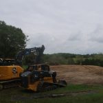 Two John Deere construction vehicles, a bulldozer and an excavator, are parked on grass beside a large, freshly excavated area for building a pole barn. Trees surround the site under a cloudy sky. Kentucky Builders And Excavating