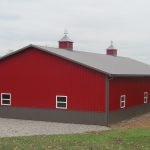 A large red pole barn with a gray roof and multiple white-trimmed windows sits on a gravel foundation in a rural setting, surrounded by trees and an overcast sky in the background. Kentucky Builders And Excavating