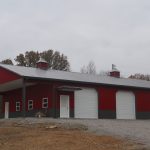 Large red and white pole barn with four garage doors, a side entrance door, and a gray metal roof, situated on a gravel lot with trees in the background under an overcast sky—ideal for building or excavating projects. Kentucky Builders And Excavating