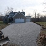 A modern two-story house with a three-car garage and a newly laid gravel driveway. A small excavating vehicle is parked to the left, and leafless trees surround the house on a cloudy day. Kentucky Builders And Excavating