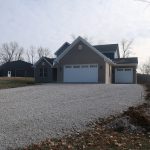 A modern house with three garage doors and a gravel driveway sits next to a pole barn, surrounded by leafless trees and a grassy yard under a cloudy sky. Kentucky Builders And Excavating