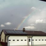 A faint rainbow arches above a large pole barn with solar panels on its roof, set against a cloudy sky and open, grassy fields. Kentucky Builders And Excavating