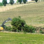 A yellow excavator is excavating near green bushes in a grassy, rural field with rolling hills and scattered trees in the background, preparing the site for a future pole barn. Kentucky Builders And Excavating