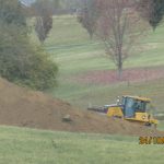 A yellow bulldozer is excavating dirt near a large soil pile in a grassy, fenced field with trees in autumn foliage. The site appears to be prepared for building, possibly a pole barn. Photo timestamp: 24/10/2023 20:57. Kentucky Builders And Excavating