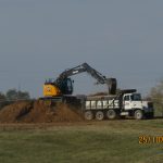 An excavator is excavating dirt into a white dump truck on a grassy field, with a house and trees in the background—possibly preparing for a building or pole barn. The date and time stamp on the photo is 25/10/2023 01:49. Kentucky Builders And Excavating