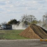 A dump truck drives on a dirt road, raising dust behind it. In the foreground, there’s a large pile of dirt and part of an excavator—excavating for a building project—with trees in the background. The date stamp reads 25/10/2023 01:52. Kentucky Builders And Excavating