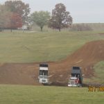 Two dump trucks with raised beds unload dirt on a grassy field, preparing the ground for building or excavating a pole barn. Piles of soil, trees, and a house are visible in the background. The date and time 27/10/2023 19:26 appear on the image. Kentucky Builders And Excavating