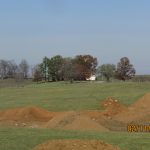 A grassy landscape with mounds of dirt from recent excavating in the foreground, scattered trees, and a white house in the distance under a clear blue sky. The date 02/11/2023 and time 23:22 are stamped on the image. Kentucky Builders And Excavating