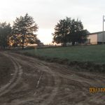 A dirt road curves through grassy land toward a group of trees and a pole barn at sunset. The sky glows orange, and the uneven ground shows tire tracks from recent excavating. The date on the photo reads 03/11/2023, 04:29. Kentucky Builders And Excavating