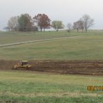 Three construction vehicles—an excavator excavating dirt into a truck, and two bulldozers—work on a large grassy field, preparing the site for building a pole barn, with trees and a fence in the background on a cloudy day. Kentucky Builders And Excavating