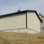 A beige metal pole barn or shed with a black roof sits on a raised concrete foundation beside a beige house. The bare soil in front suggests recent excavating, while the mostly cloudy sky looms overhead. Kentucky Builders And Excavating