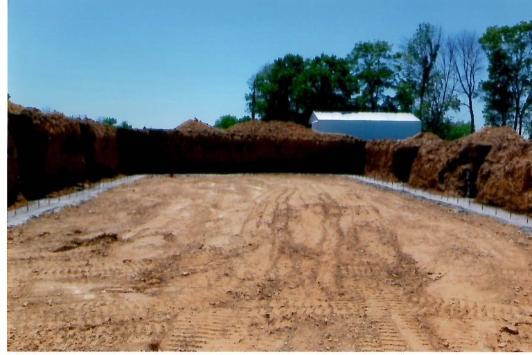 A large, rectangular construction site with dirt ground and high piles of soil from recent excavating; trees and a white building are visible in the background under a clear blue sky. Kentucky Builders And Excavating