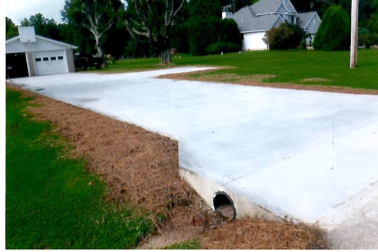 A concrete driveway with a drainage pipe underneath, created after excavating, runs through a grassy yard toward a house and garage, which sit amid trees and greenery, with space nearby perfect for building a pole barn. Kentucky Builders And Excavating