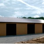 A large, tan metal pole barn with four wide open garage doors sits on a gravel surface, surrounded by trees and a blue sky with clouds in the background. Kentucky Builders And Excavating