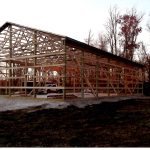 A wooden frame of a large pole barn under construction stands on a concrete foundation, with a pickup truck parked inside. Leafless trees surround the building, and the ground is partly covered in grass. Kentucky Builders And Excavating