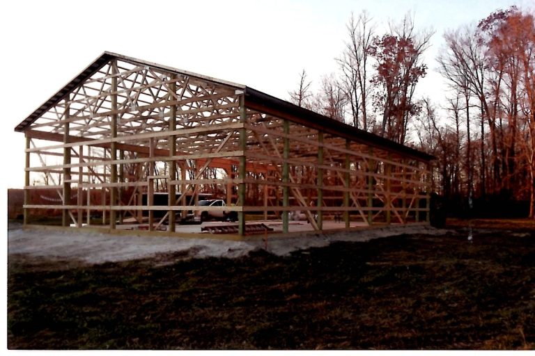 A wooden frame of a large pole barn under construction stands on a concrete foundation, with a pickup truck parked inside. Leafless trees surround the building, and the ground is partly covered in grass. Kentucky Builders And Excavating