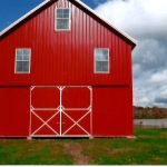 A bright red pole barn with white trim sits on green grass under a partly cloudy sky. The barn has four white-trimmed windows, large double doors in front, and stands proudly among trees and a pond visible in the background. Kentucky Builders And Excavating