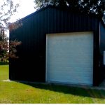 A black metal pole barn garage with a large white roll-up door and four windows on the side sits on a grassy lawn with trees and a curved driveway in the background. Kentucky Builders And Excavating