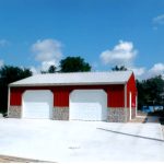 A red metal pole barn garage with two white roll-up doors sits on a spacious, clean concrete driveway. The building features a gray metal roof, stone accents at the base, and is surrounded by trees under a partly cloudy sky. Kentucky Builders And Excavating