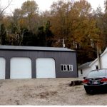 A gray three-door pole barn garage sits on a dirt lot next to a white house; a black car and an ATV are parked nearby, with autumn trees in the background. Kentucky Builders And Excavating
