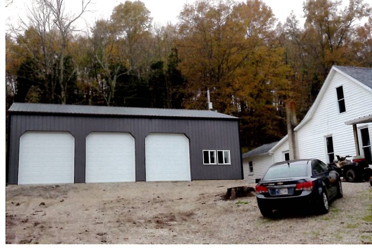 A gray three-door pole barn garage sits on a dirt lot next to a white house; a black car and an ATV are parked nearby, with autumn trees in the background. Kentucky Builders And Excavating