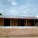 A large metal pole barn building with a brown roof and four open bays stands on a gravel and dirt lot. Trees and greenery surround the building, while a small vehicle, possibly used for excavating, is parked on the right. The sky is partly cloudy. Kentucky Builders And Excavating