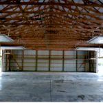 Interior view of a spacious empty pole barn with a high ceiling, exposed wooden beams, concrete floor, and open sliding doors letting in natural light from the sides. Kentucky Builders And Excavating