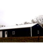 A dark blue metal pole barn with a white roof, white trim, a small window, and a white garage door stands on a grassy area with trees and a cloudy sky in the background, perfect for building projects or excavating equipment storage. Kentucky Builders And Excavating