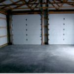 Interior of an empty pole barn garage with a concrete floor, wooden beams, and two closed white garage doors. The space is clean and well-lit, with visible ceiling supports—ideal for building projects or storing equipment after excavating work. Kentucky Builders And Excavating