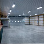 A spacious pole barn or warehouse with concrete floors, white paneled walls, exposed wooden beams, and bright overhead lights. Multiple garage doors and a small loft area on the left side complete this versatile building. Kentucky Builders And Excavating