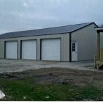 A large, light gray metal pole barn garage with three white roll-up doors and a side entrance sits next to a deck attached to a house, on gravel and patchy grass under a cloudy sky—ideal for building projects or excavating equipment storage. Kentucky Builders And Excavating