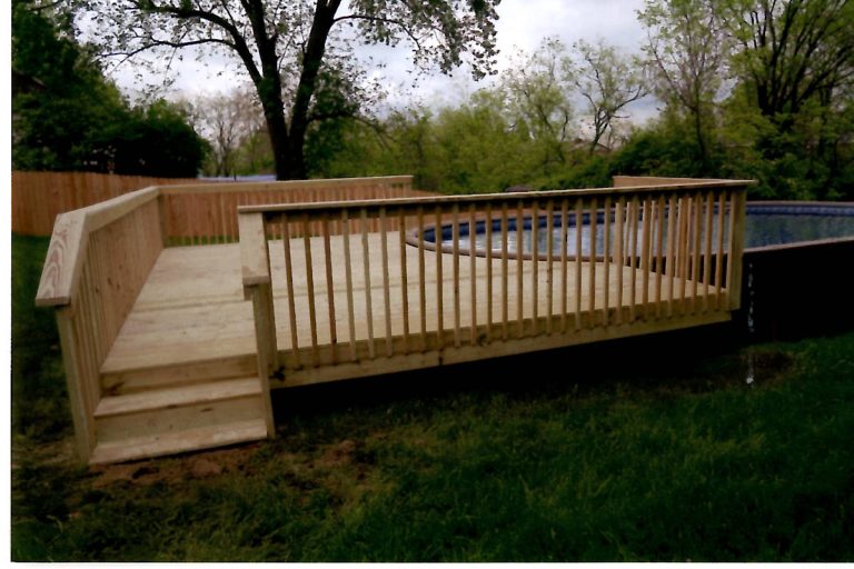 A wooden deck with railing and stairs leads to a round above-ground pool in a grassy backyard surrounded by trees, a wooden fence, and a nearby pole barn. Kentucky Builders And Excavating
