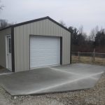 A small, light gray metal building with a white roll-up door and side entry door sits on a newly poured concrete driveway, the result of recent excavating work. Its surrounded by gravel, a chain-link fence, with trees and grass in the background. Kentucky Builders And Excavating