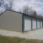 A large metal pole barn garage with four white roll-up doors stands on a gravel driveway, surrounded by leafless trees and grass under a partly cloudy sky. Kentucky Builders And Excavating