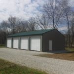 A large metal pole barn with a green roof and four white roll-up doors sits on a gravel driveway, surrounded by leafless trees and grass under a partly cloudy sky. Kentucky Builders And Excavating