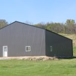 A large gray metal building with a white door and window sits on a grassy plot, surrounded by trees and a clear blue sky in the background. Kentucky Builders And Excavating